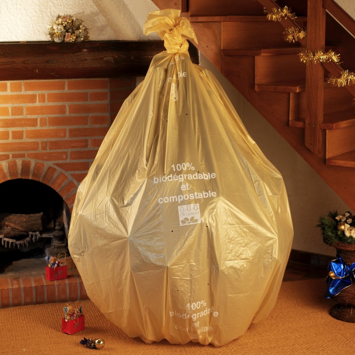 Grand sac poubelle doré, rempli, posé près d'une cheminée en briques, ambiance festive.