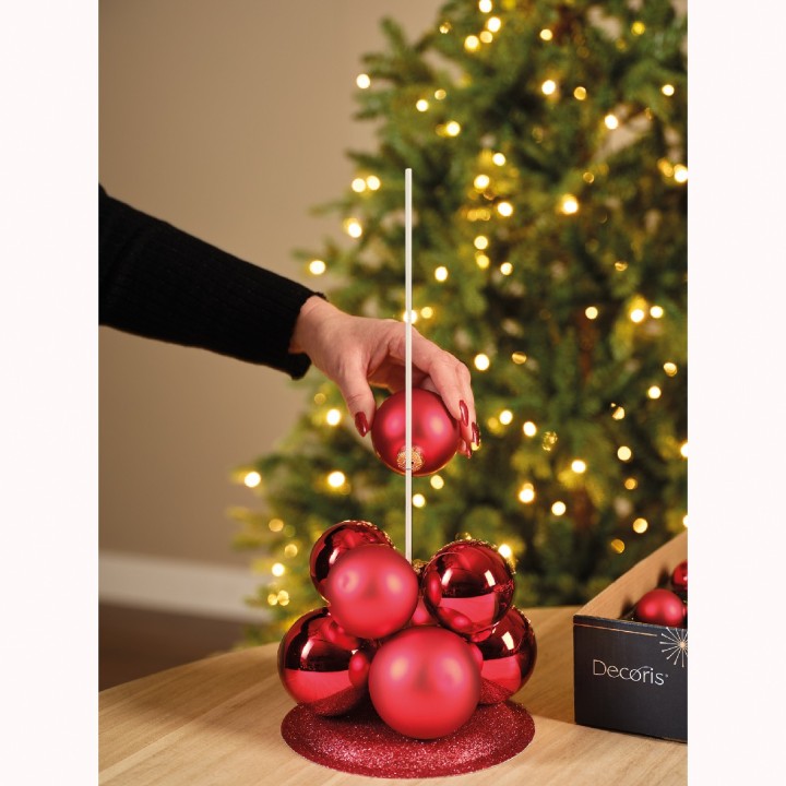 Main plaçant des boules rouges brillantes sur une base scintillante, devant un sapin illuminé.