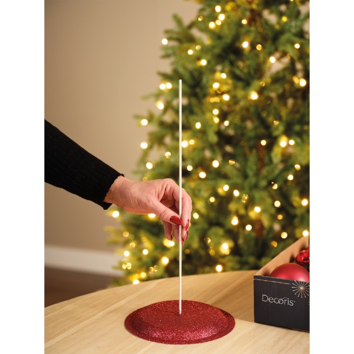 Une main place un bâton blanc sur une base rouge scintillante près d'un sapin illuminé.