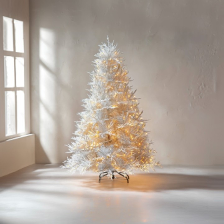 Sapin artificiel blanc illuminé, posé sur un sol clair, devant une fenêtre baignée de lumière.